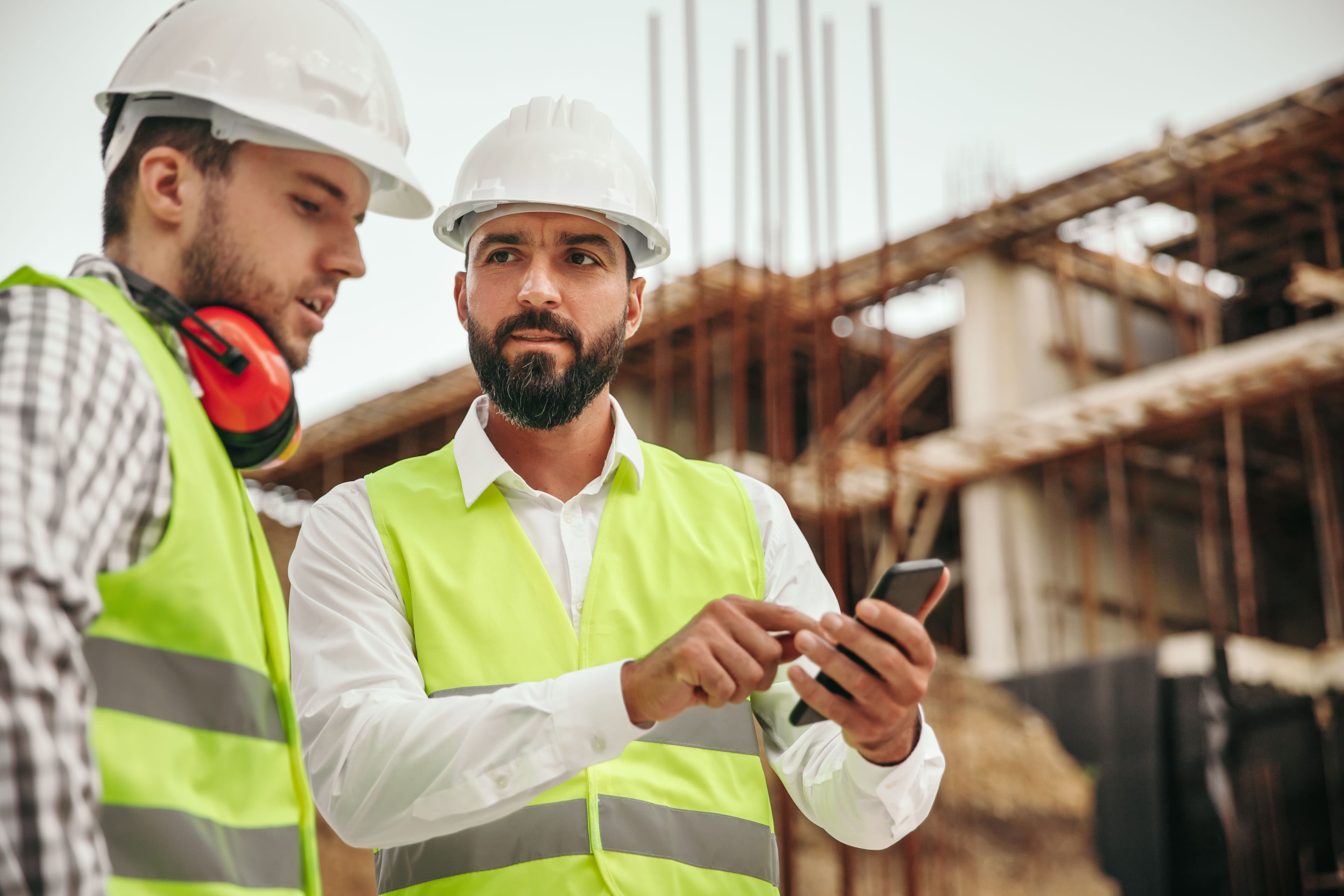 Two construction workers stood on a building site, both looking down at a mobile phone that one of the construction workers is holding.